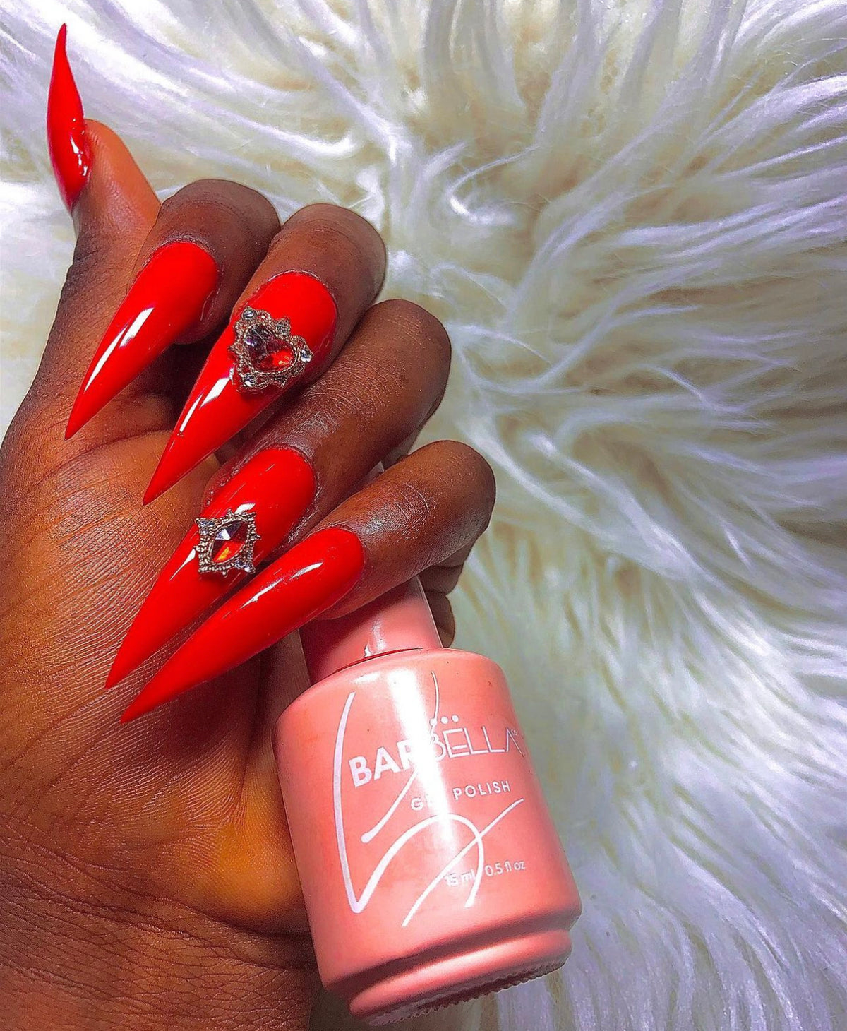 Hand with long bright red stiletto nails and heart-shaped rhinestone accents holding a peach BarBella Co. polish bottle against white faux fur.