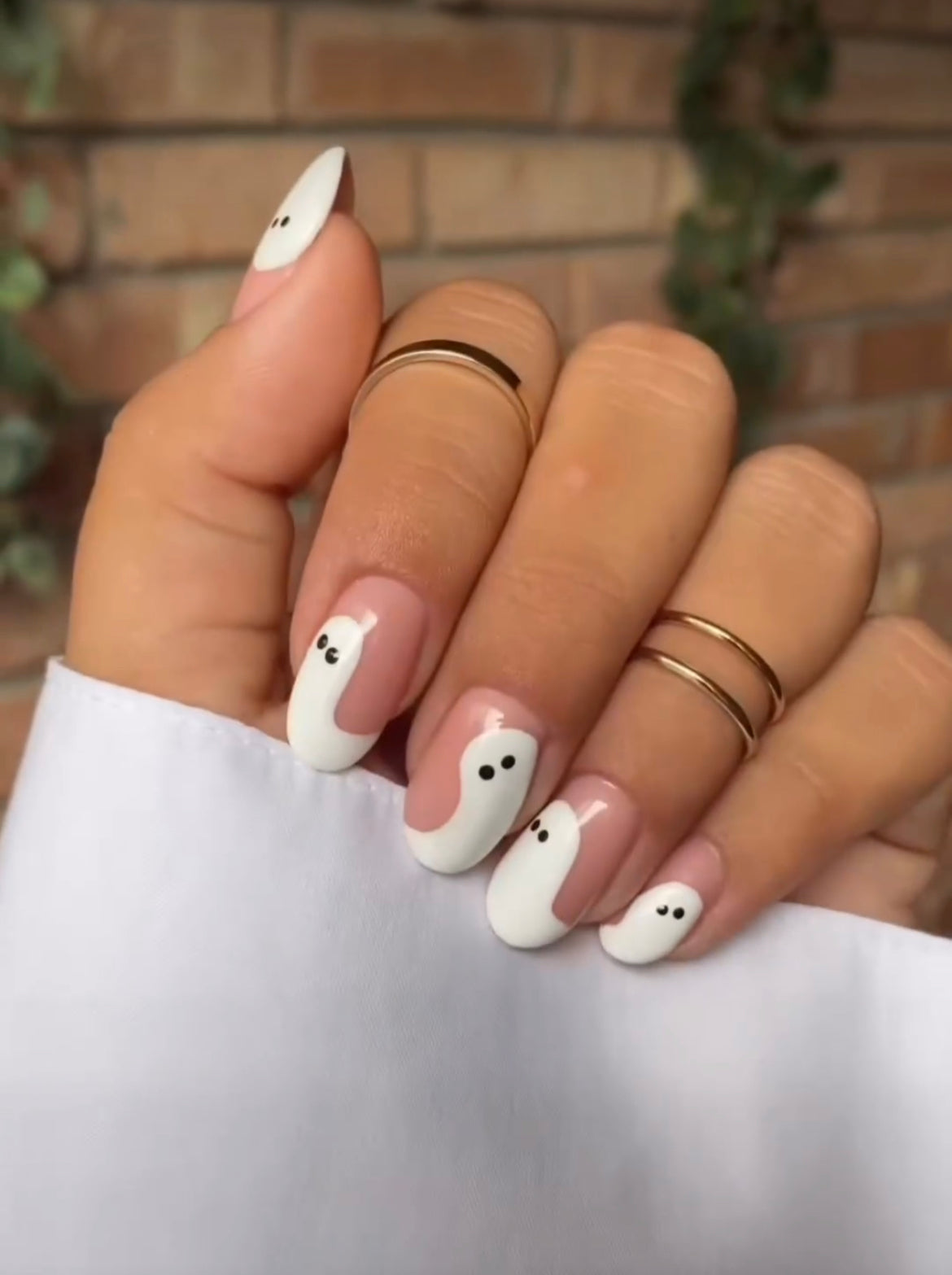 Hand with nude almond nails painted with white ghosts with black eyes, wearing thin gold rings, against a brick wall. Minimalist Halloween ghost nail art.