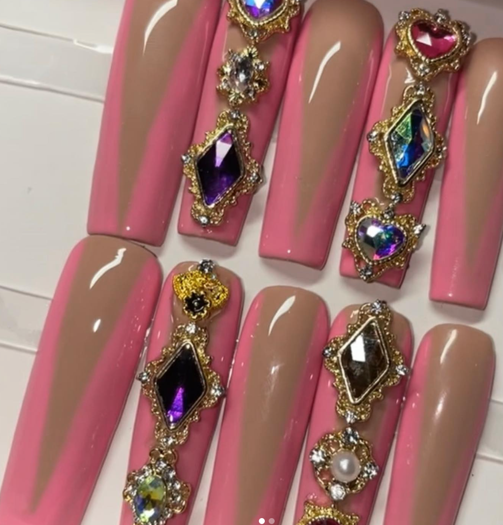 Long coffin nails in pink and nude with a V design, decorated with large gold-framed gemstone charms in purple, blue, pink and clear. Statement nail jewelry trend.